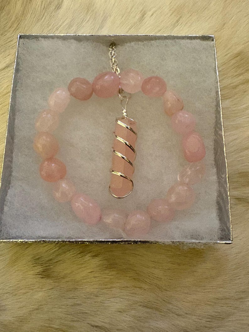 May include: A pink rose quartz bracelet and pendant necklace set. The bracelet is made of rounded pink stones. The pendant is a pink rose quartz crystal wrapped in silver wire, suspended from a silver chain. The jewelry is presented in a silver box.