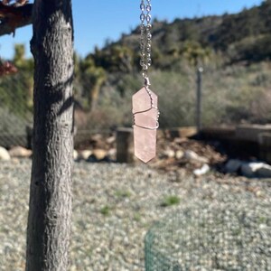 May include: A rose quartz crystal pendant necklace. The light pink crystal is wire-wrapped and suspended from a silver chain. The necklace hangs against a backdrop of a tree, blue sky, and a hillside.