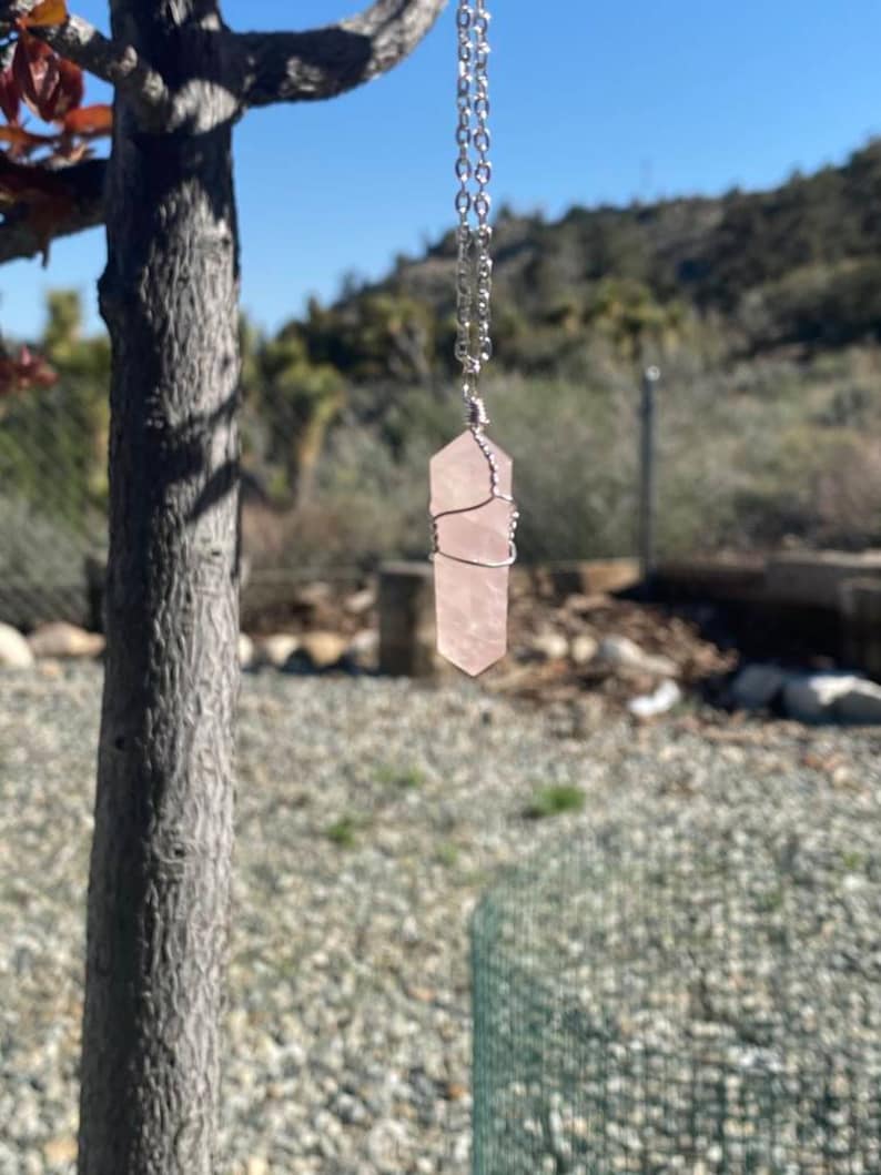 May include: A rose quartz crystal pendant necklace. The light pink crystal is wire-wrapped and suspended from a silver chain. The necklace hangs against a backdrop of a tree, blue sky, and a hillside.