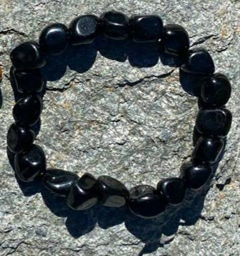 May include: A black gemstone bracelet made with smooth, rounded beads. The bracelet is laid out on a grey, textured surface.