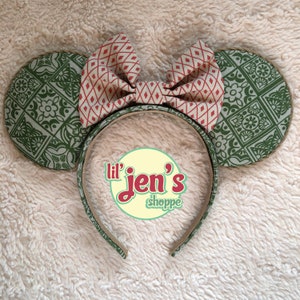 May include: Green and red patterned fabric mouse ears with a brown headband and a bow. The headband has the text "lil' jen's shoppe" on it.