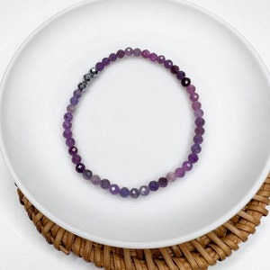 May include: A delicate bracelet featuring faceted purple beads with a gradient effect, transitioning from deep violet to lighter shades. Accented with a few metallic gray beads. The bracelet is displayed on a white plate and a woven coaster.
