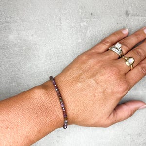 May include: A beaded bracelet with small, faceted beads in shades of purple, green, and brown. The bracelet is worn on a wrist, showcasing the delicate design and color variations. The bracelet is a piece of jewelry.