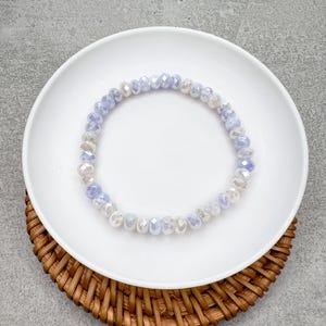 May include: A delicate bracelet made of iridescent glass beads in shades of lavender and white. The beads are faceted and create a shimmering effect.