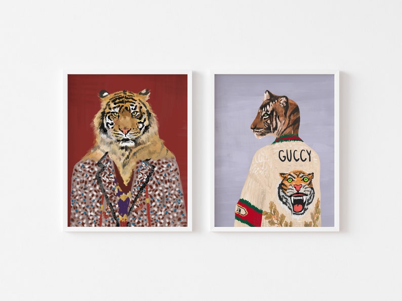 May include: Two framed illustrations of tigers wearing clothing. The tiger on the left is wearing a brown and black patterned jacket with a red background. The tiger on the right is wearing a white sweater with a green and red Gucci logo and a tiger graphic.