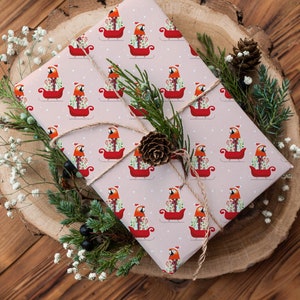 May include: A pink gift wrap with a repeating pattern of red cardinals riding in sleighs. The gift is wrapped with twine and decorated with small white flowers and pine sprigs.