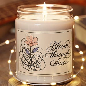 Könnte beinhalten: Eine weiße Kerze in einem Glasgefäß mit einem Etikett, auf dem "Bloom through chaos" steht. Das Etikett zeigt auch eine stilisierte Blume mit rosa Blütenblättern und blauen Blättern. Die Kerze steht auf einem Holzuntersetzer und ist von weißen Lichterketten umgeben.