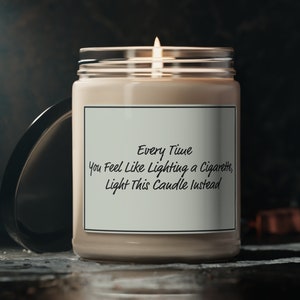 May include: A beige candle with a glass jar and a white label that reads "Every Time You Feel Like Lighting a Cigarette, Light This Candle Instead". The candle is lit and the flame is visible.