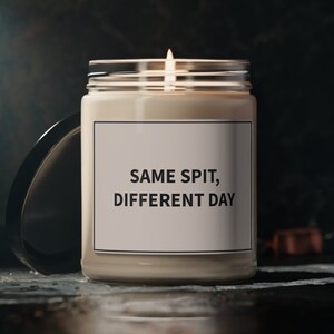 May include: A white candle with a black label that reads "SAME SPIT, DIFFERENT DAY". The candle is lit and sitting on a dark surface.
