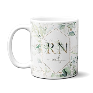 May include: White ceramic mug with a gold hexagonal frame and the letters "RN" in gold. The frame is surrounded by a watercolor design of green leaves and branches. The name "emily" is written in cursive below the letters.