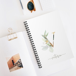 May include: A white spiral bound notebook with a gold and green watercolor letter V and the word "victoria" written in cursive.