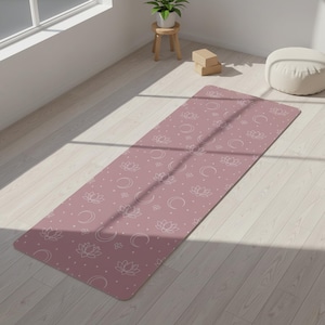 May include: A rectangular yoga mat in a dusty rose color featuring a repeating white line art pattern of lotus flowers, crescent moons, and small dots. The mat is laid out flat on a light-colored wooden floor, displaying a consistent design.