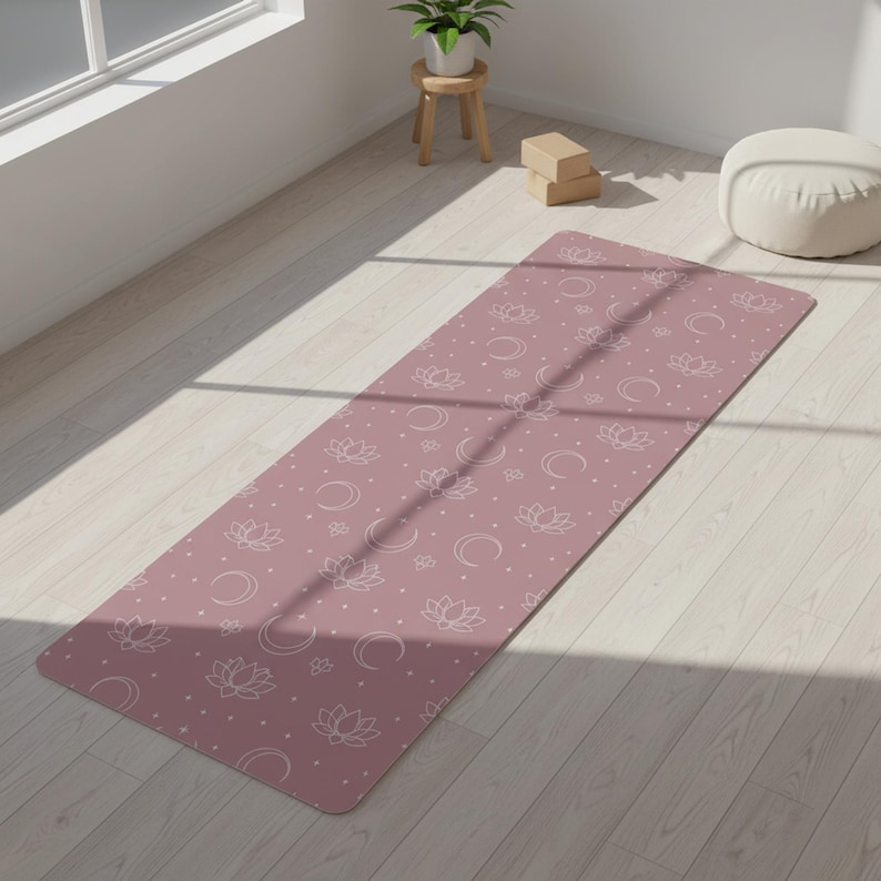 May include: A rectangular yoga mat in a dusty rose color featuring a repeating white line art pattern of lotus flowers, crescent moons, and small dots. The mat is laid out flat on a light-colored wooden floor, displaying a consistent design.