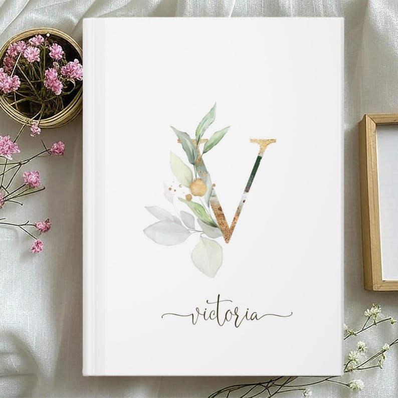 Pu&ograve; includere: Un diario bianco con il nome "Victoria" in corsivo. La lettera "V" &egrave; decorata con foglie acquerellate dorate e verdi e dettagli dorati. Un piccolo cestino di fiori rosa &egrave; sullo sfondo.