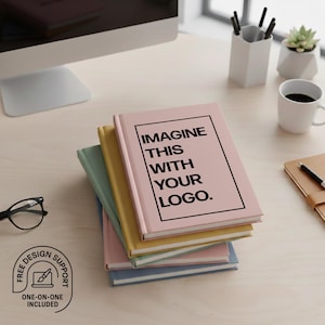 May include: A stack of notebooks in various colors, including pink, yellow, and blue, are arranged on a light wood surface. The top notebook is pink and features the text "IMAGINE THIS WITH YOUR LOGO." A pair of glasses and a coffee cup are also visible.