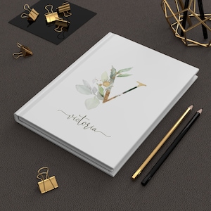 Pu&ograve; includere: Diario con copertina rigida bianca con il nome "Victoria" in corsivo. La copertina presenta un design floreale ad acquerello con la lettera "V" dorata. Matite dorate e nere e fermagli dorati su una superficie grigio scuro.