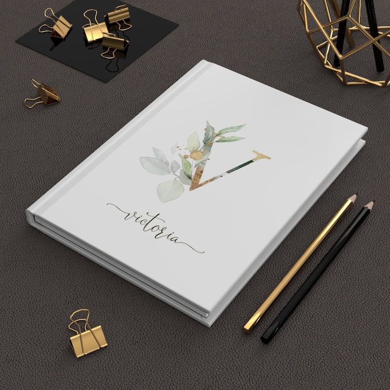 Pu&ograve; includere: Diario con copertina rigida bianca con il nome "Victoria" in corsivo. La copertina presenta un design floreale ad acquerello con la lettera "V" dorata. Matite dorate e nere e fermagli dorati su una superficie grigio scuro.