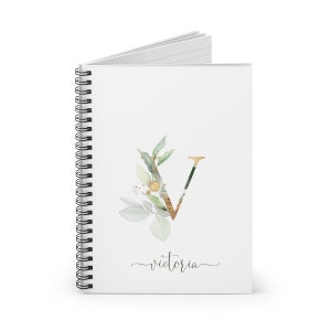 Pu&ograve; includere: Quaderno a spirale bianco con un design floreale oro e verde e la lettera "V". La parola "Victoria" &egrave; scritta in corsivo sotto il disegno floreale. Il quaderno &egrave; aperto.