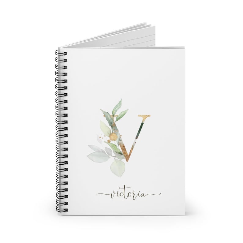 Pu&ograve; includere: Quaderno a spirale bianco con un design floreale oro e verde e la lettera "V". La parola "Victoria" &egrave; scritta in corsivo sotto il disegno floreale. Il quaderno &egrave; aperto.