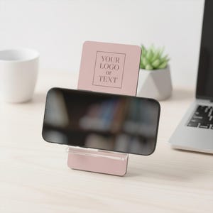 May include: A blush pink phone stand with a clear plastic base, holding a black smartphone. The stand features a vertical panel with the text "YOUR LOGO OR TEXT" in a square frame. A white mug, a succulent, and a laptop are in the background.