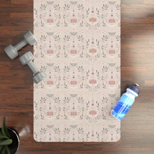 May include: A rectangular yoga mat featuring a repeating pattern of pink lotus flowers, grey leafy branches, and moon phases on a light beige background, accompanied by two grey dumbbells and a blue water bottle with a white cap.