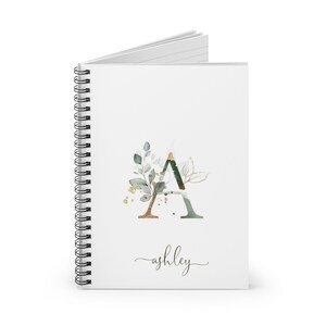 May include: A white spiral bound notebook with a green and gold watercolor letter A with leaves and gold accents. The name "ashley" is written in cursive below the letter.