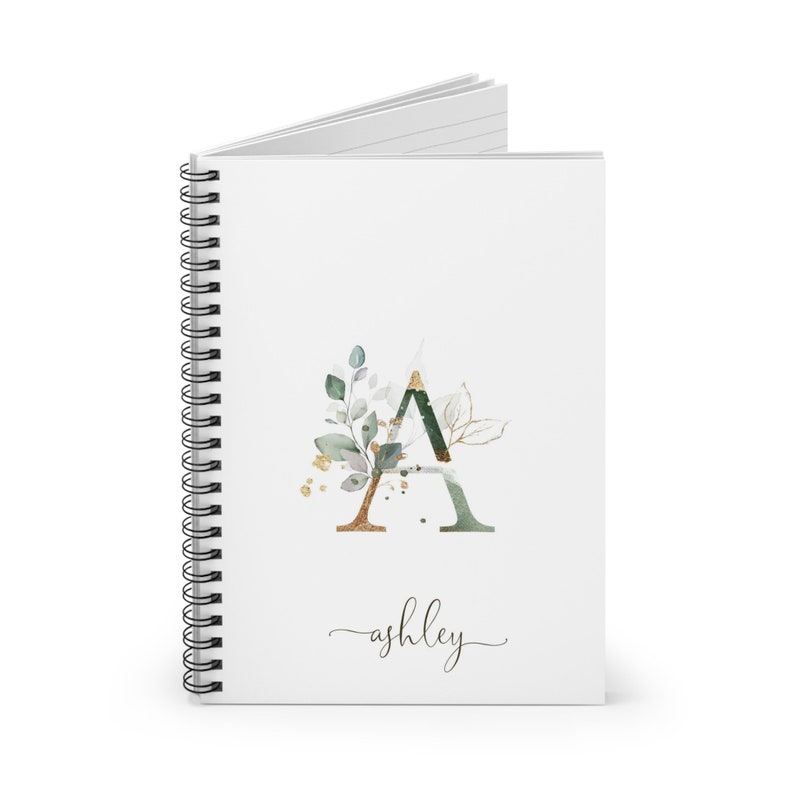 May include: A white spiral bound notebook with a green and gold watercolor letter A with leaves and gold accents. The name "ashley" is written in cursive below the letter.