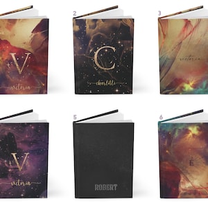 May include: Six different notebook covers with a galaxy theme. The covers feature a variety of colors, including gold, black, purple, and red. Each cover has a different initial and name printed on it, including Victoria, Charlotte, Robert, and Emma.