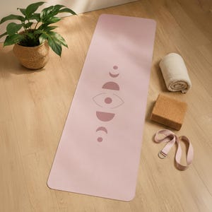 May include: A rectangular light pink yoga mat featuring a centered dark pink graphic of moon phases and an eye symbol. The mat is accompanied by a rolled white towel, a rectangular brown cork yoga block, and a light pink yoga strap with a metal ring.