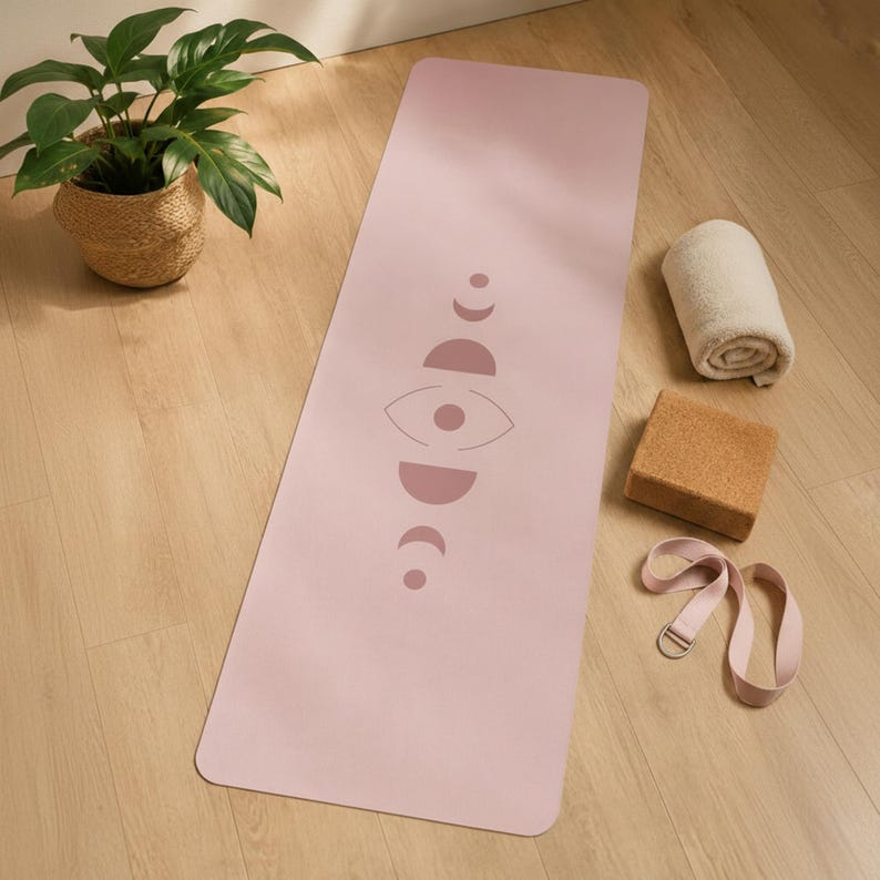 May include: A rectangular light pink yoga mat featuring a centered dark pink graphic of moon phases and an eye symbol. The mat is accompanied by a rolled white towel, a rectangular brown cork yoga block, and a light pink yoga strap with a metal ring.