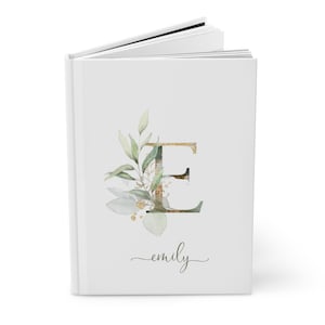 Pu&ograve; includere: Un diario bianco con la lettera "E" dorata e verde, ornato da foglie e elementi floreali ad acquerello. Il nome "emily" &egrave; scritto in corsivo sotto il disegno. Il diario &egrave; aperto, rivelando pagine bianche.
