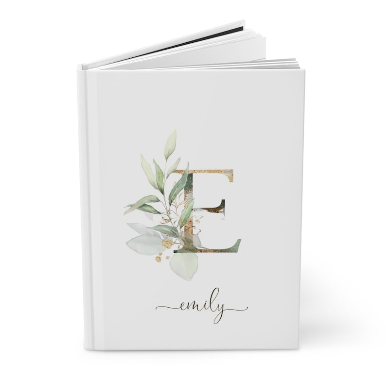 Pu&ograve; includere: Un diario bianco con la lettera "E" dorata e verde, ornato da foglie e elementi floreali ad acquerello. Il nome "emily" &egrave; scritto in corsivo sotto il disegno. Il diario &egrave; aperto, rivelando pagine bianche.