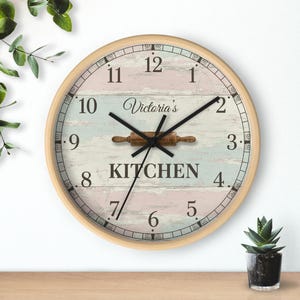 Könnte beinhalten: Runde Küchenuhr mit hellem Holzrahmen. Das Zifferblatt hat ein verwittertes Pastellmuster mit der Aufschrift "Victoria's Kitchen" und einer Teigrolle. Schwarze Zeiger zeigen die Uhrzeit an.