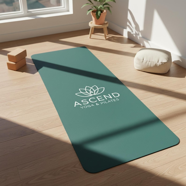 May include: A rectangular teal yoga mat featuring a white lotus flower graphic centered above the text ASCEND YOGA & PILATES. The mat has rounded corners and a smooth surface, positioned flat on a light-colored wooden floor surface.