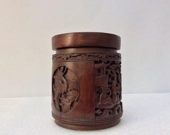 May include: A cylindrical, dark brown wooden container with intricate carvings. The container features detailed relief carvings of figures and scenes, possibly depicting a historical or cultural narrative. The top and base are smooth, while the body is adorned with the carved designs.