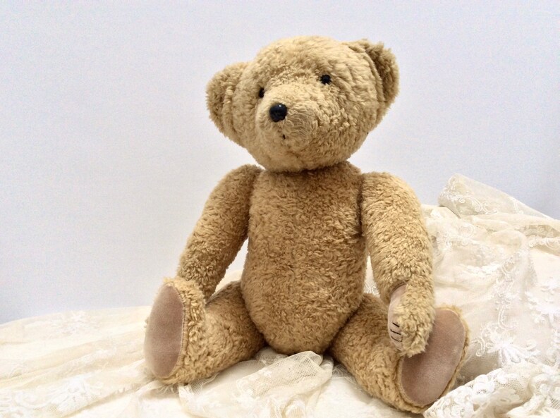 May include: A plush brown teddy bear sitting on a white lace fabric. The bear has a soft, fuzzy texture and black button eyes.