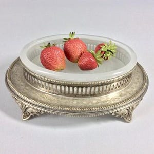 Puede incluir: Una fuente plateada ornamentada con un inserto de vidrio transparente, que contiene varias fresas frescas. La fuente tiene un diseño detallado en relieve y descansa sobre pequeños pies. Las fresas son rojas con tallos verdes.