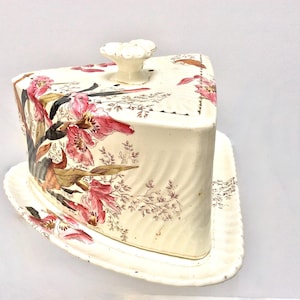 May include: A white ceramic cheese dome with a floral design in shades of pink, brown, and green. The dome has a lid with a knob on top. The dome sits on a matching plate.