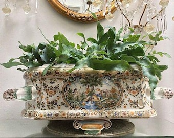 Op de afbeelding: Een rechthoekige, antieke keramische plantenbak gevuld met groen gebladerte. De plantenbak heeft een gedetailleerd bloemen- en landschapsontwerp in blauw, geel en bruin. Het heeft twee handvatten aan de zijkant en staat op een decoratieve basis.