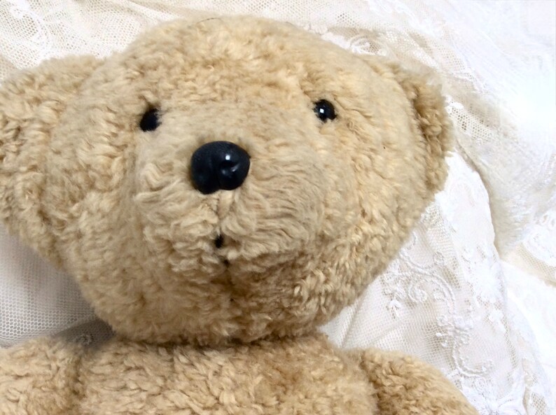 May include: A close-up of a beige teddy bear with black eyes and a black nose. The bear is lying on a white lace fabric.