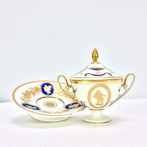 May include: A white porcelain sugar bowl and saucer with gold and blue accents. The bowl has a lid with a gold cherub design and the saucer has a gold floral design.