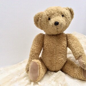 May include: A plush brown teddy bear sitting on a white lace fabric. The bear has black eyes and a black nose.