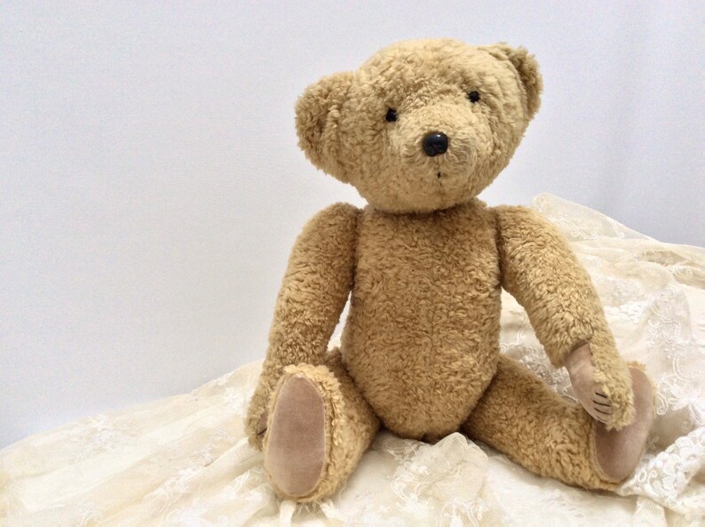 May include: A plush brown teddy bear sitting on a white lace fabric. The bear has black eyes and a black nose.