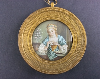 Könnte beinhalten: Ein runder, antik goldfarbener Rahmen umgibt ein Miniaturgemälde einer Frau in einem blau-gelben Kleid. Das Gemälde zeigt eine Frau, die eine Tasse hält, mit einer Topfpflanze im Hintergrund.