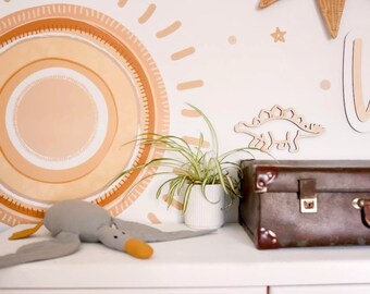 May include: A white wall with a large sun graphic in shades of orange and brown. The sun graphic has a concentric circle design. There are small stars and dots scattered around the sun. A small potted plant sits in front of the sun graphic. A grey stuffed toy duck is lying on a white surface. A brown suitcase is on the right side of the image.