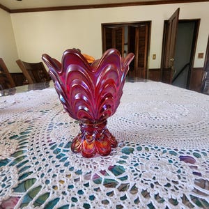 May include: A red iridescent glass bowl with a scalloped rim and a pedestal base. The bowl is sitting on a white crocheted tablecloth.