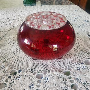 May include: A round, red glass flower frog with a clear, grid-like top. The red glass has a textured appearance. The frog sits on a white, crocheted tablecloth. A green glass cake stand is visible in the background.