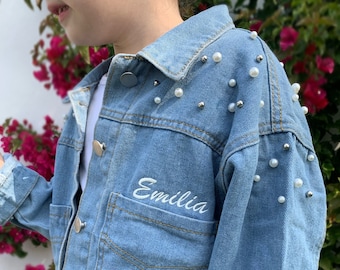 Könnte beinhalten: Eine hellblaue Jeansjacke mit Perlenbesatz an den Schultern und dem Namen "Emilia" auf der Tasche gestickt.