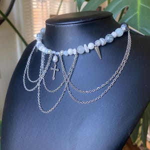 May include: A silver chain choker necklace with a white and gray stone beaded strand. The necklace features a silver cross charm and a silver spike charm.