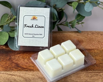 Op de afbeelding: Zes witte wasmelts in een transparante plastic bak. De bak heeft een label met de tekst "Fresh Linen 100% Natural Soy Wax Melt 3 oz."